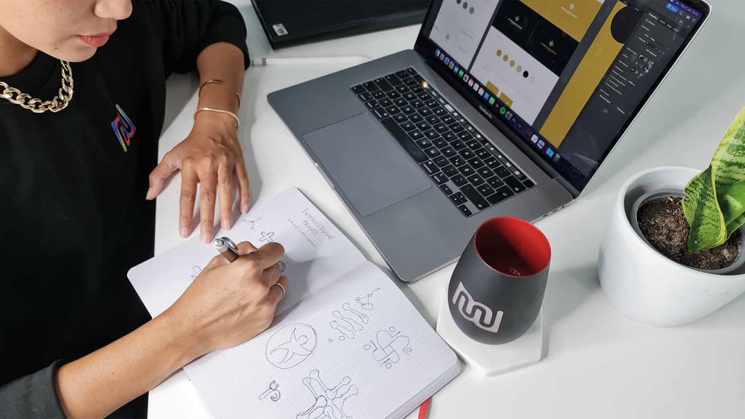 Designerin skizziert Logo-Entwürfe auf Papier, daneben Laptop und Tasse mit Mischler-Logo